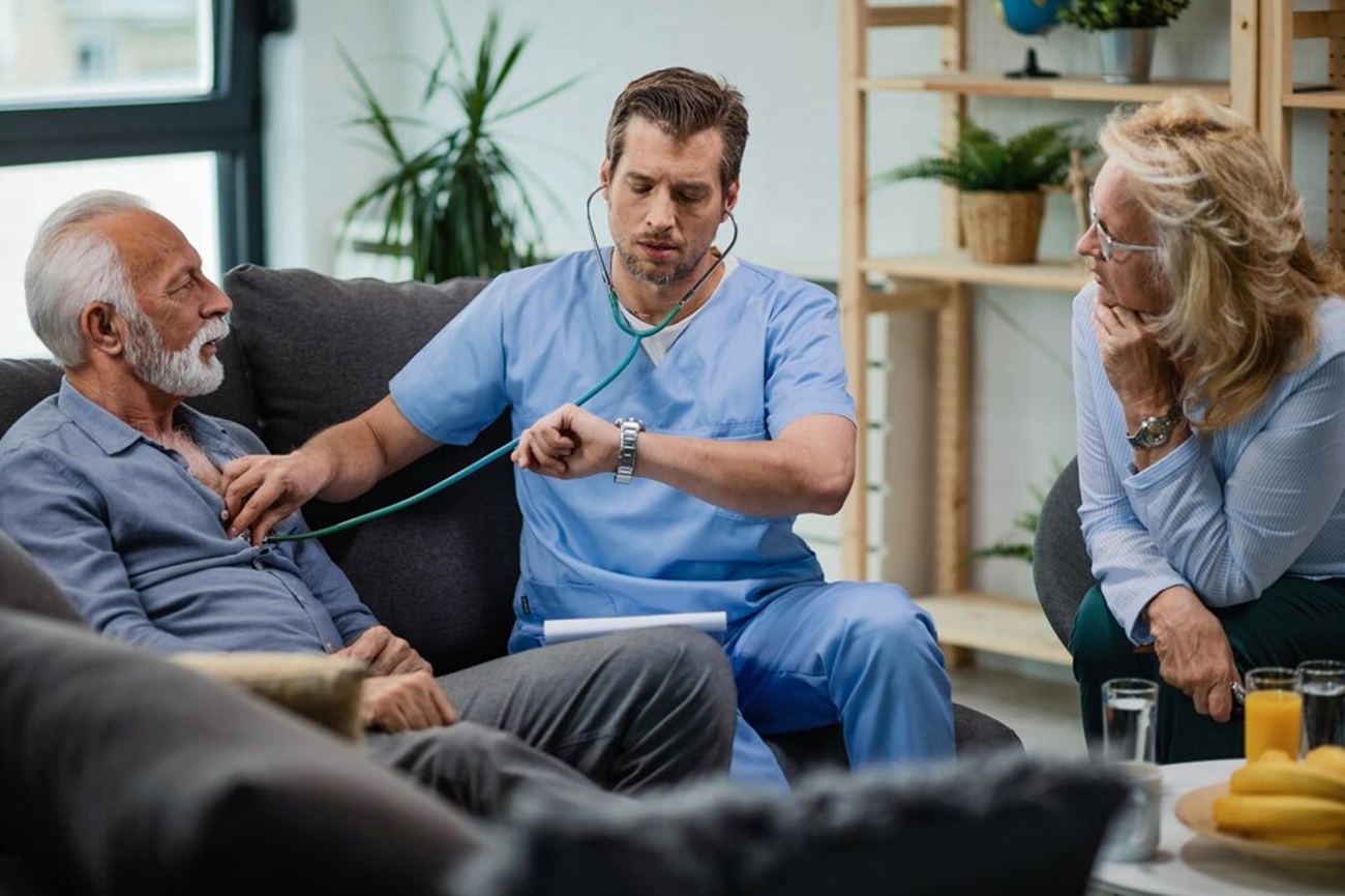 doctor-visiting-senior-couple-home-checking-his-wristwatch-while-listening-heartbeat-man_637285-1385